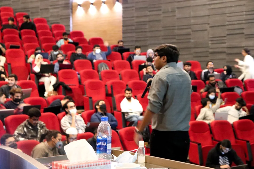 A full university auditorium of attendees with laptops open, seen from the front of the room