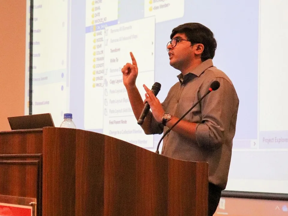 Speaking at a lectern with a microphone, gesturing mid-sentence, a data modelling tool projected behind